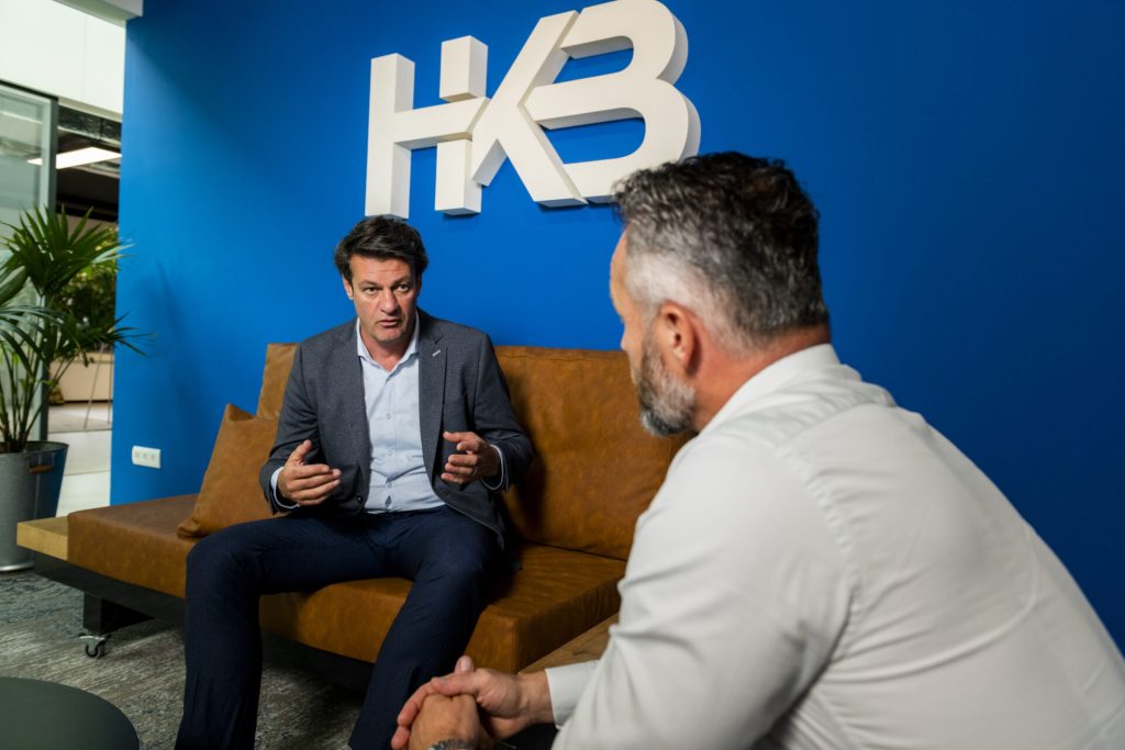 Twee mannen praten in kantoor met blauw logo.