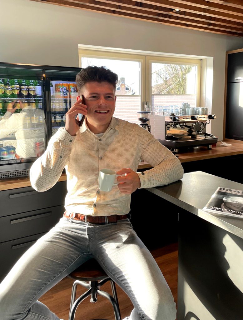 Man telefoneert in café met koffiemachine op achtergrond.