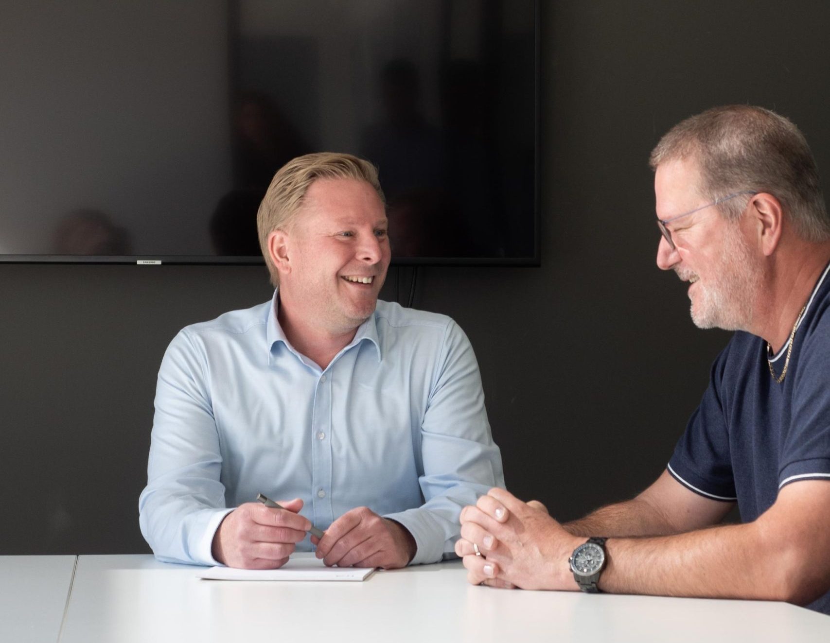 Twee mannen in gesprek bij vergadertafel.