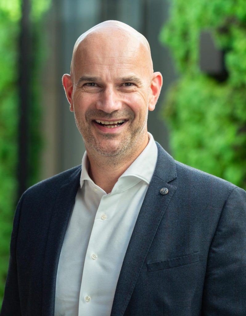 Glimlachende man in formele kleding, groene achtergrond.