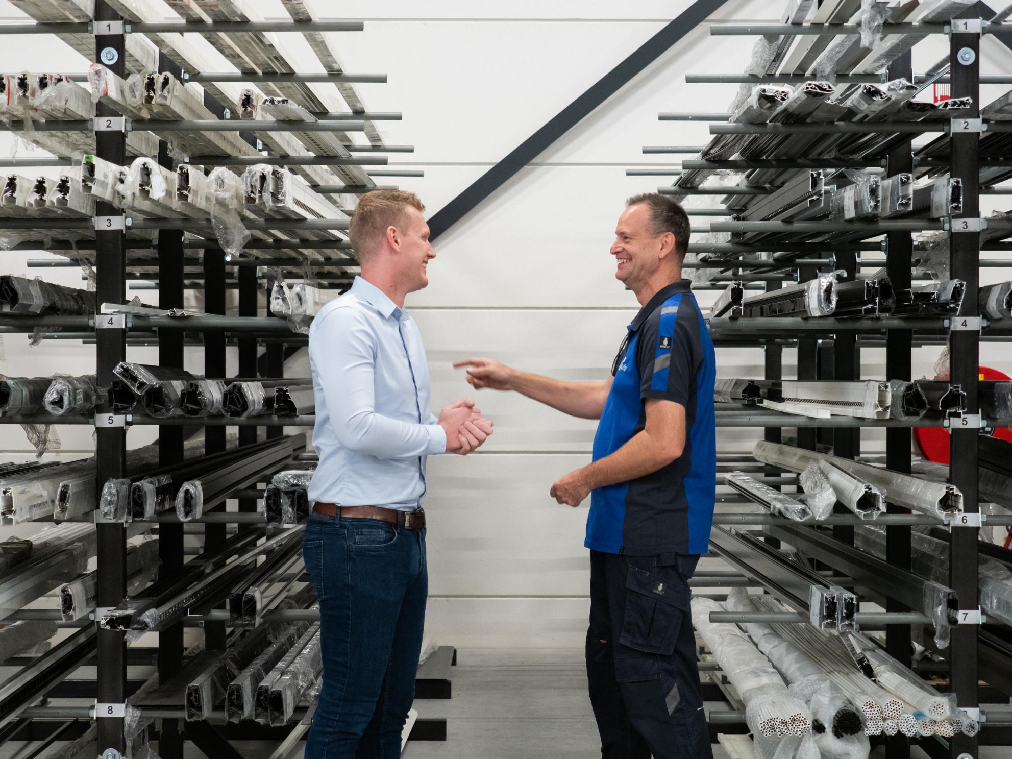 Twee mannen praten in fabriek met stalen rekken.