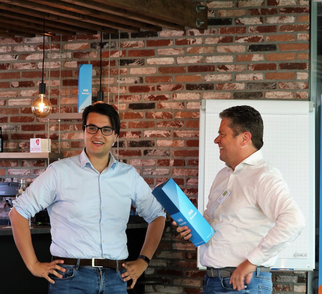 Twee mannen lachen en praten voor een bakstenen muur.