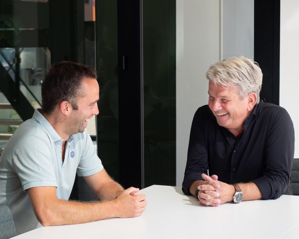 Twee mannen lachen en praten aan tafel.