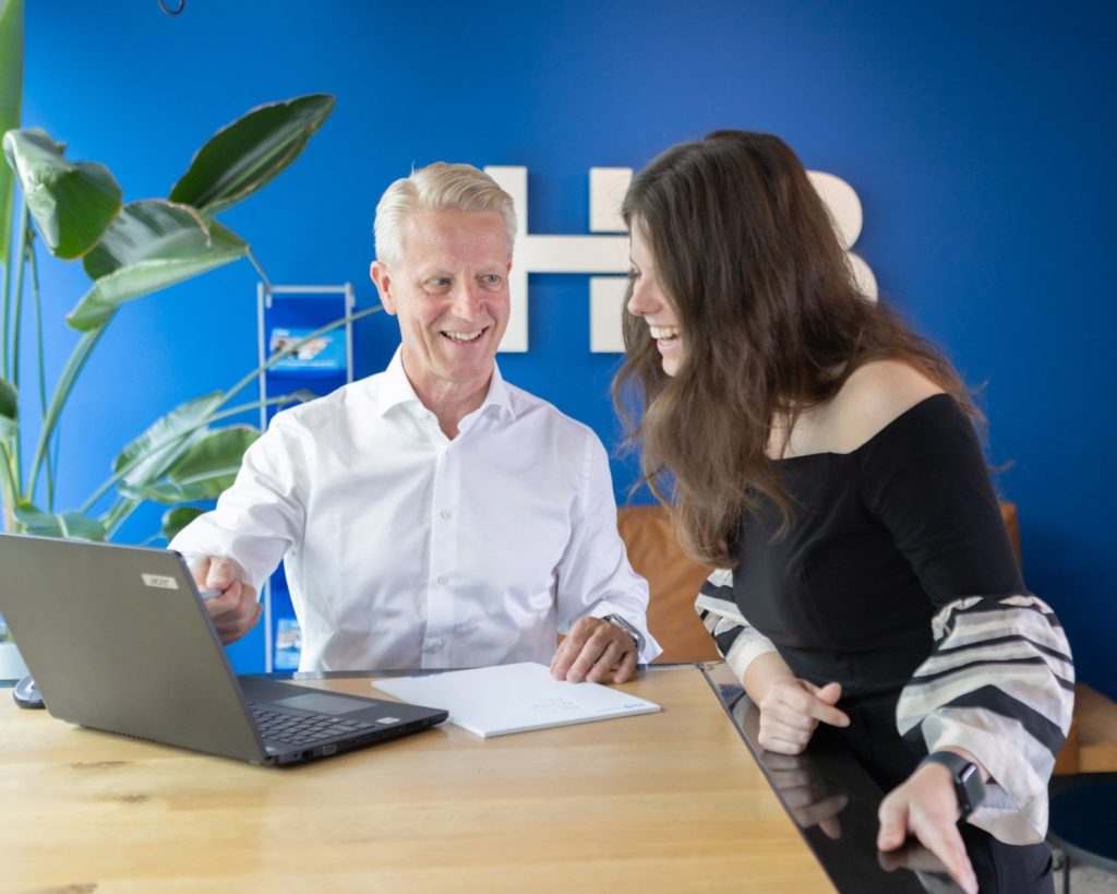 Twee mensen lachen bij laptop in kantooromgeving.