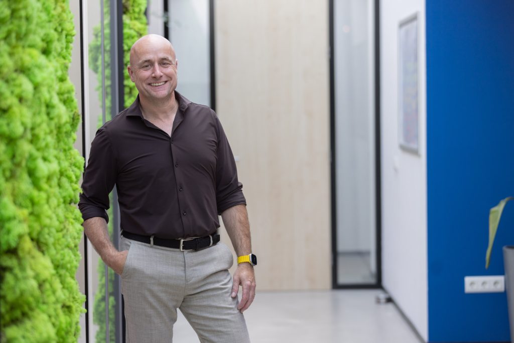 Man in kantoor naast groene muur glimlacht.